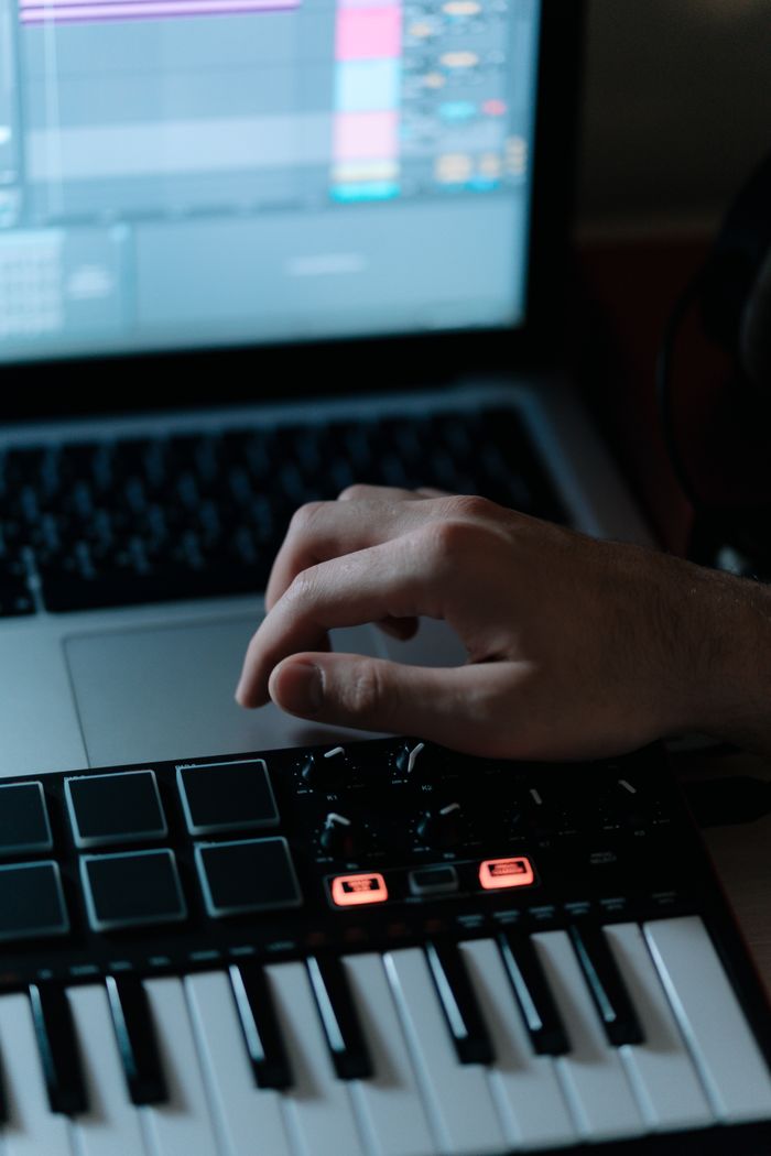 About Close-up of hand adjusting synthesizer controls with laptop screen displaying audio software.